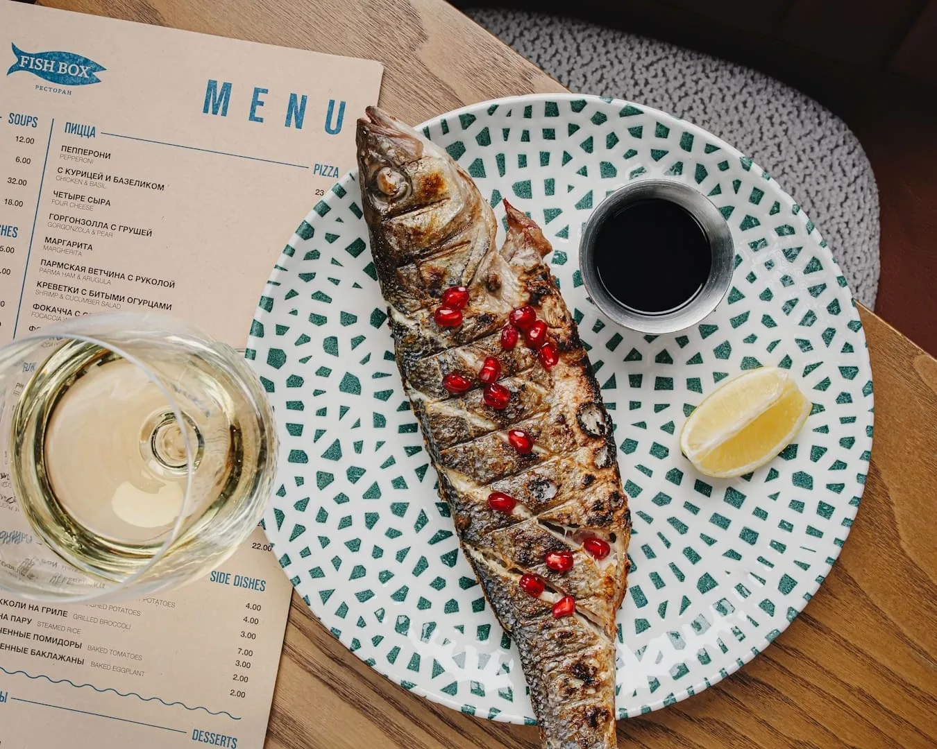 Рыба на гриле с гранатом, рядом меню Fish Box и белое вино